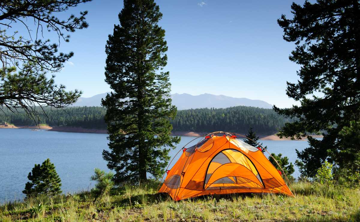An orange tent setup on a grassy hill overlooking a beautiful pristine blue lake, surrounded by pine trees.