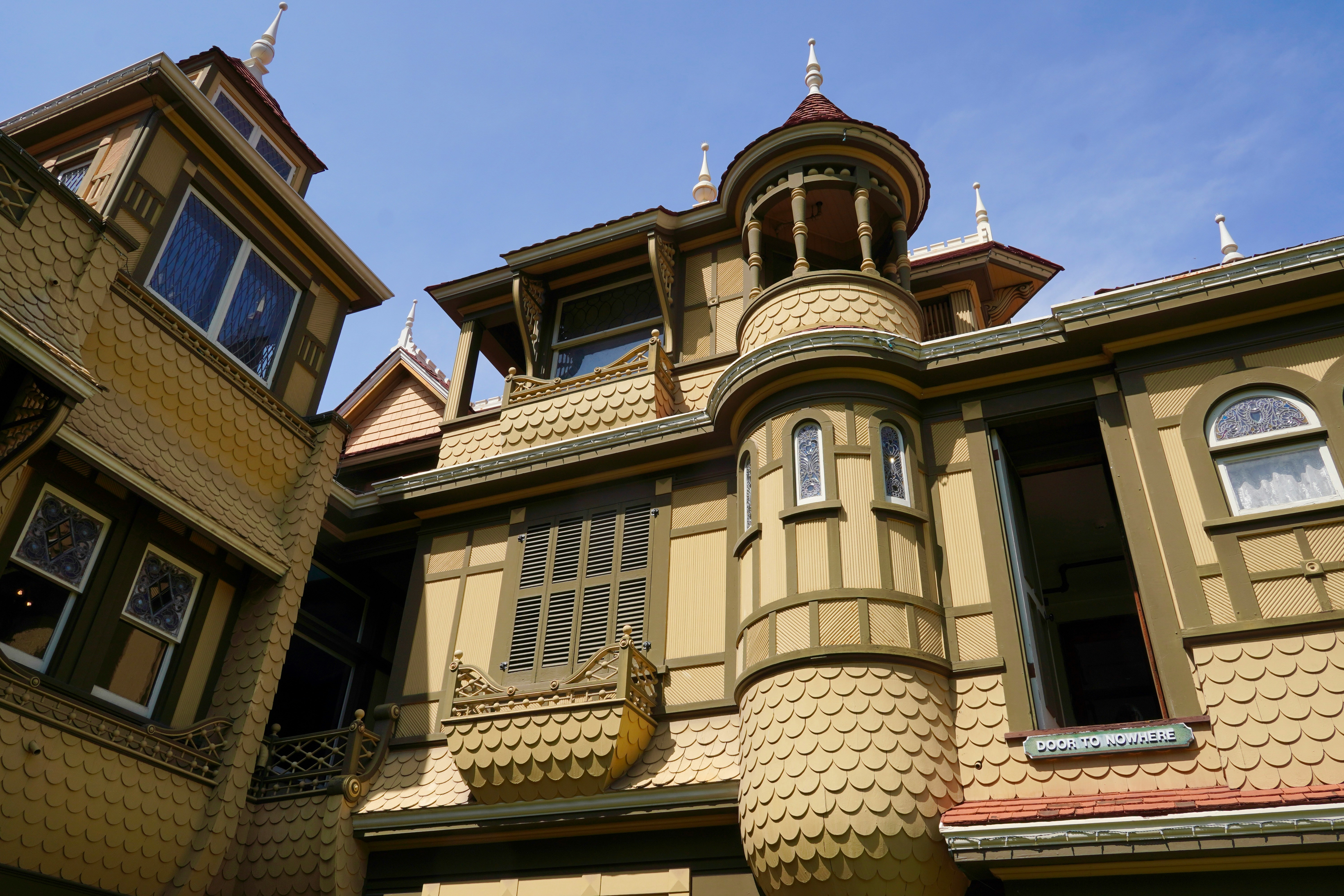 An ornate turret and textured exterior walls of the Winchester Mystery House showcase intricate Victorian architecture under a clear blue sky. Stained glass windows and decorative trim add to the surreal beauty, while a sign reading "Door to Nowhere" marks a famously strange and haunting feature of the mansion. The whimsical design hints at the home's mysterious history and eccentric construction.